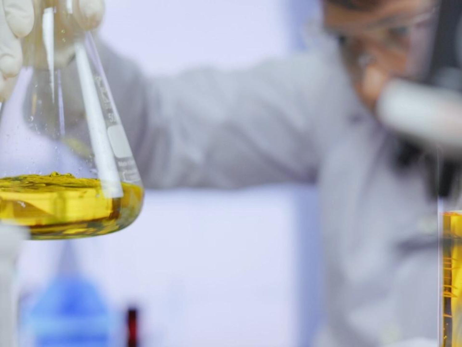 A scientist in a laboratory examines a yellow chemical solution in a glass flask.